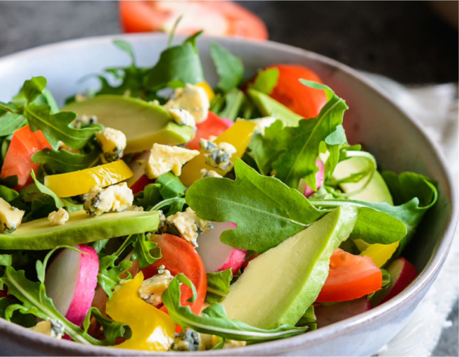 A close-up of a fresh vegetable salad in a gray bowl, featuring sliced tomatoes, cucumbers, yellow bell peppers, radishes, arugula, and crumbled blue cheese.